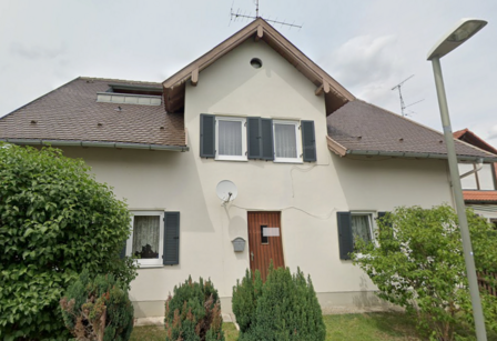 Vorderansicht eines Hauses mit Satteldach, Fensterläden, Holzhaustür und Sträuchern