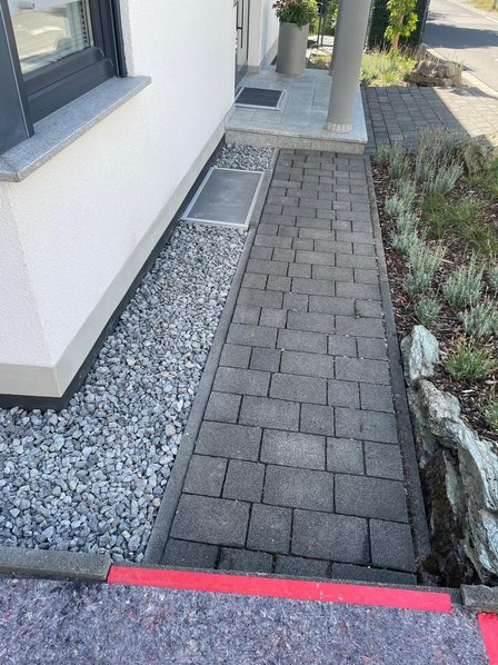 Gepflasterter Gehweg neben Hausfassade mit Kiesstreifen und Lichtschachtabdeckung