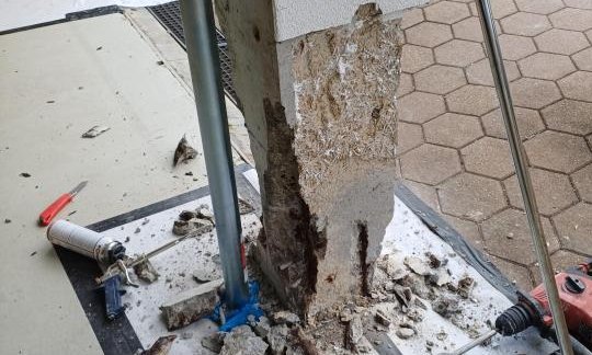 Abgeplatzter Sockelbereich an Stuetze, Bauschutt und Bohrhammer auf Fliesenboden.