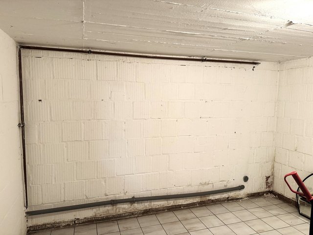 Kellerraum mit weiß beschichteter Wand, dunkle Verfärbungen am Wandsockel und graue Rohrleitung