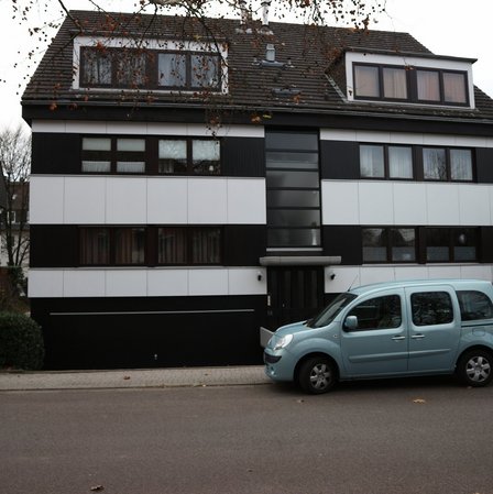 Mehrstöckiges Wohnhaus mit dunkler Fassade und weißen Paneelen, Auto am Straßenrand
