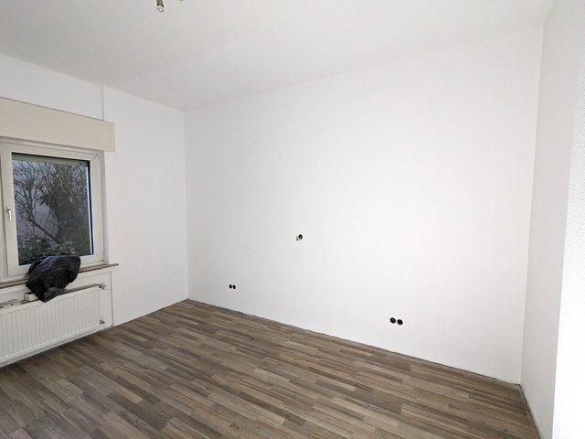 Leerer Raum mit weißer Wand, Fenster und Heizkörper, Steckdosenöffnungen sichtbar.