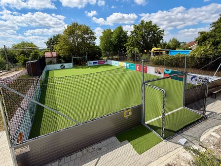 Eingezäunter Kunstrasen-Sportplatz mit Bandenwerbung, Bäumen und blauem Himmel.