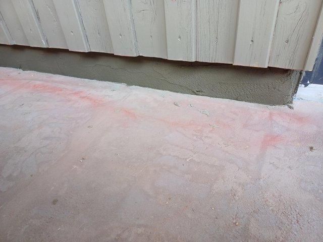 Graue Masse auf Wandsockel unter grau gestrichener Holzverschalung, Boden mit rötlichen Markierungen