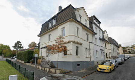 Mehrstöckiges helles Wohngebäude an Straße, parkende Autos, Herbstbaum im Vordergrund