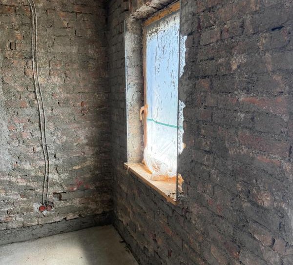 Unverputzter Ziegelraum mit Fenster, Folienabdeckung und sichtbaren Kabeln an der Ecke