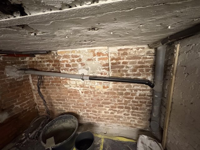 Freigelegte Ziegelwand im Keller mit Abflussrohren und niedriger Betondecke