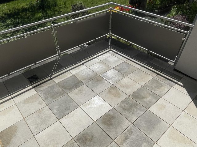 Gefliester Balkon mit Metallgeländer und grauen Sichtschutzplatten, Sonnenlicht und Schatten.
