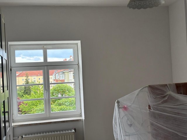 Helles Zimmer mit Fenster, Heizkoerper und abgedecktem Bett, Blick auf Baeume und Haeuser.