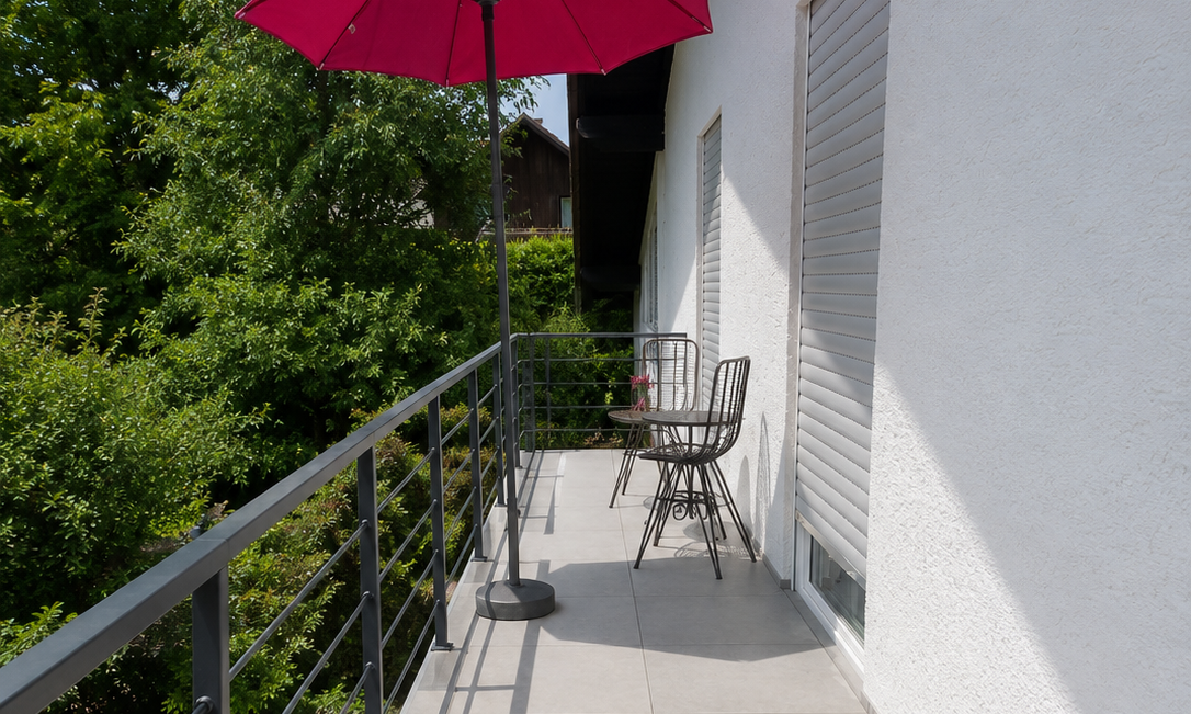 Balkon mit grauen Fliesen, Metallgeländer, Tisch und Stühlen unter pinkem Sonnenschirm