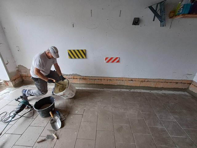 Arbeiter mischt graue Masse vor freigelegtem Wandstreifen in gefliestem Raum