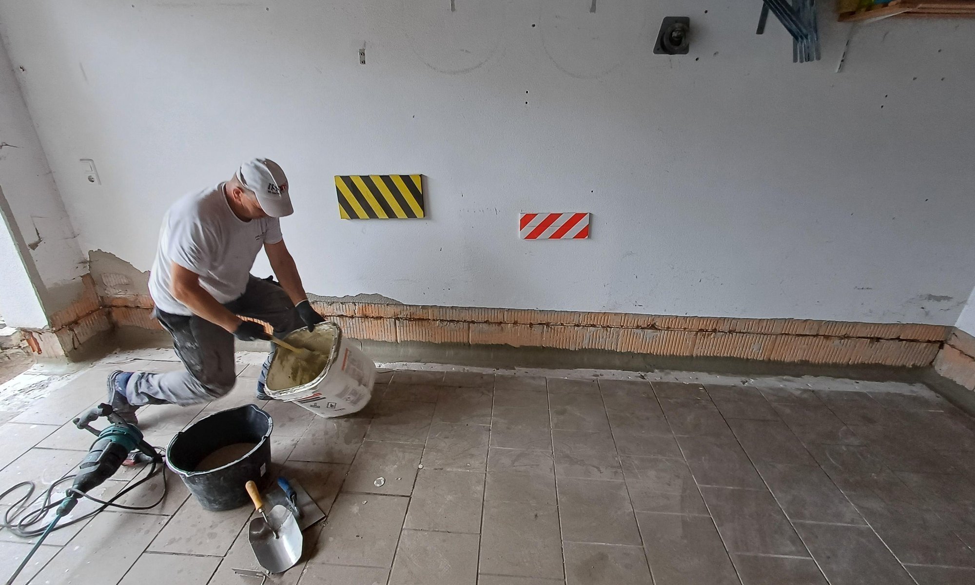 Arbeiter mischt graue Masse vor freigelegtem Wandstreifen in gefliestem Raum