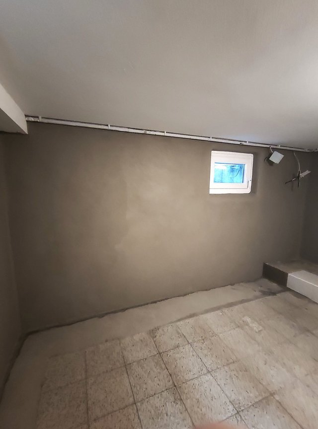 Graue Kellerwand mit kleinem Fenster, sichtbaren Leitungen und Fliesenboden.