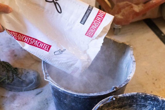 Hand schüttet weißes Pulver aus Beutel in schwarzen Eimer.