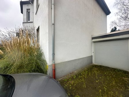 Weiße Hausfassade mit Fallrohr neben Garagentor und Bewuchs am Sockel