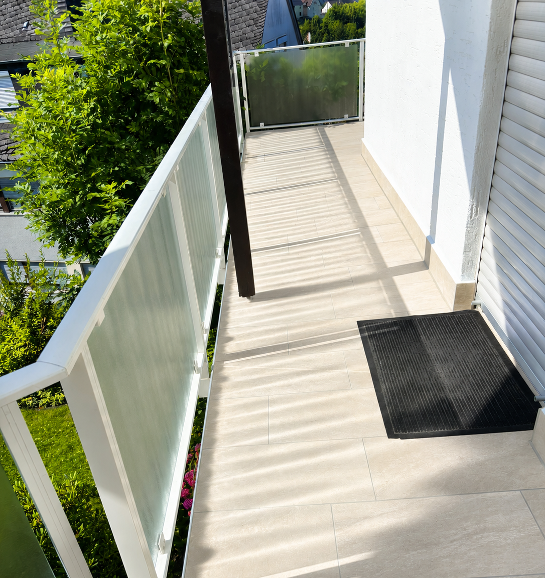 Sonniger Balkon mit beigen Bodenfliesen, Milchglasgeländer und schwarzer Fußmatte