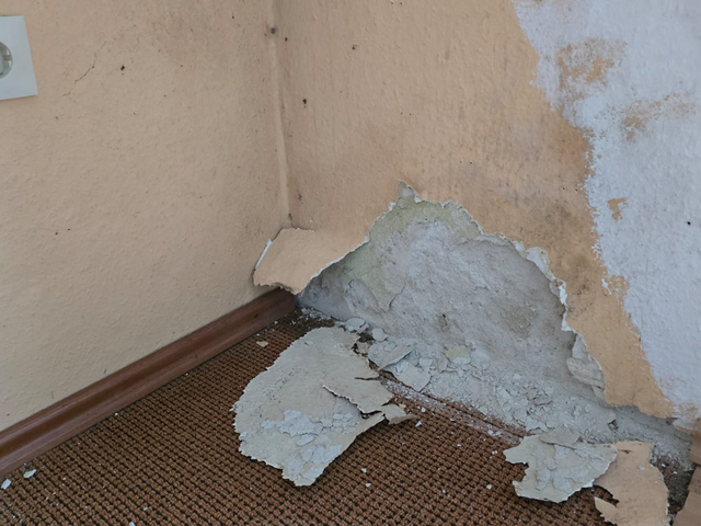 Abblätternde Beschichtung an Zimmerecke mit freiliegender hellgrauer Fläche