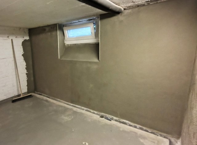 Frisch verputzte Kellerwand mit kleinem Fenster und freiliegendem Rohr an der Decke