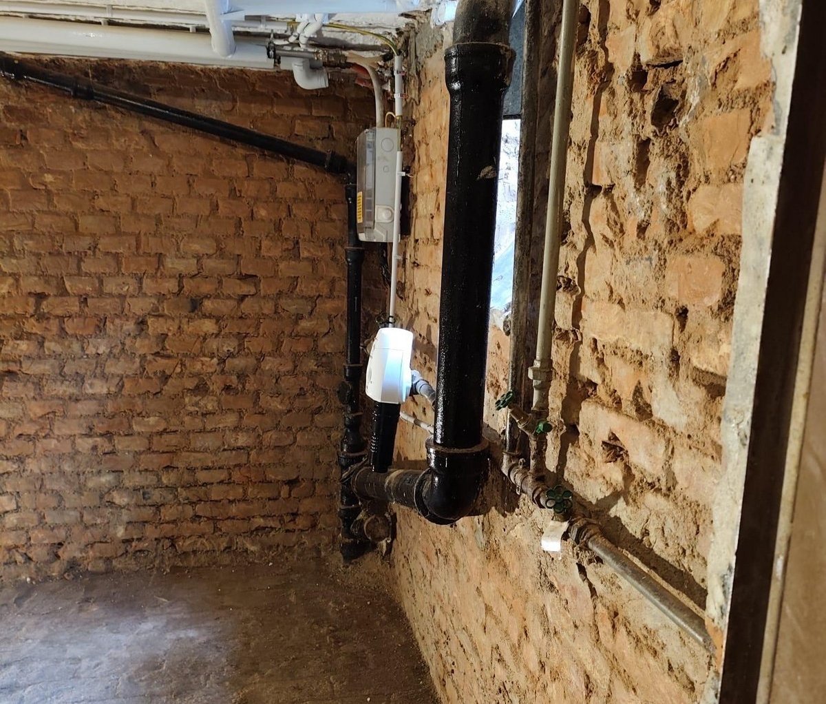 Kellerraum mit freiliegender Ziegelwand und sichtbaren Rohrleitungen an Decke und Wand
