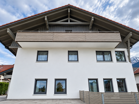 Gebäude mit weißer Fassade, Holzbalkon und dunkelgrauen Fenstern unter rotem Ziegeldach