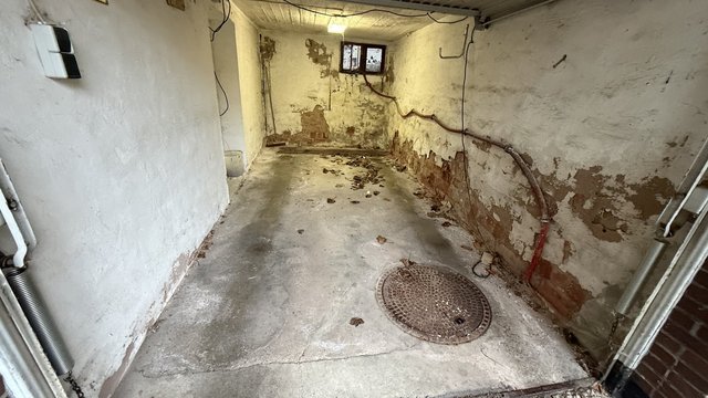 Leerer Kellerraum mit abplatzendem Putz, freigelegtem Mauerwerk und Bodenablauf