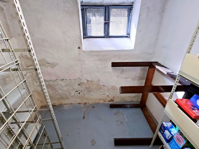 Fleckige Kellerwand mit abplatzender Beschichtung unter kleinem Gitterfenster, Regale im Raum