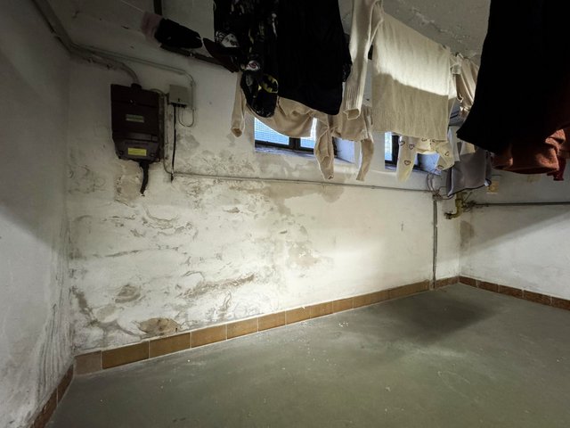 Kellerraum mit fleckiger, beschädigter Wand unter kleinen Fenstern und aufgehängter Wäsche