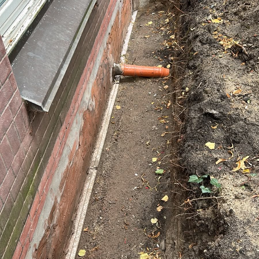 Freigelegter Erdgraben entlang einer Backstein-Kellerwand mit orangefarbenem Rohr