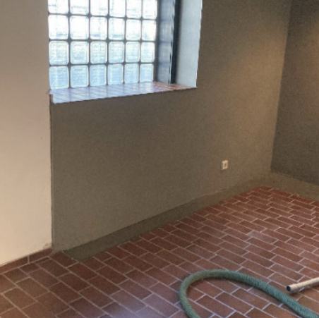 Raum mit Glasbausteinfenster, graue Wand und Terrakottafliesen, grüner Schlauch liegt auf dem Boden.