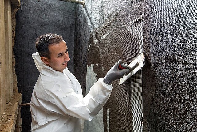 Arbeiter trägt schwarze Masse auf Kellerwand auf.
