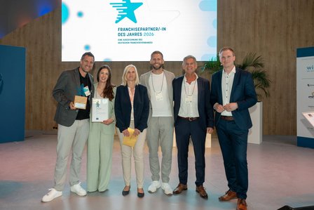 Sechs Personen posieren auf Bühne mit Trophäe und Urkunde „Franchisepartner/-in des Jahres 2026“.