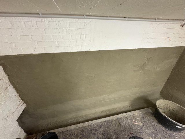 Weiße Ziegelwand über Fläche mit grauer Masse, Rohrleitung unter Decke, Mörtelkübel am Boden