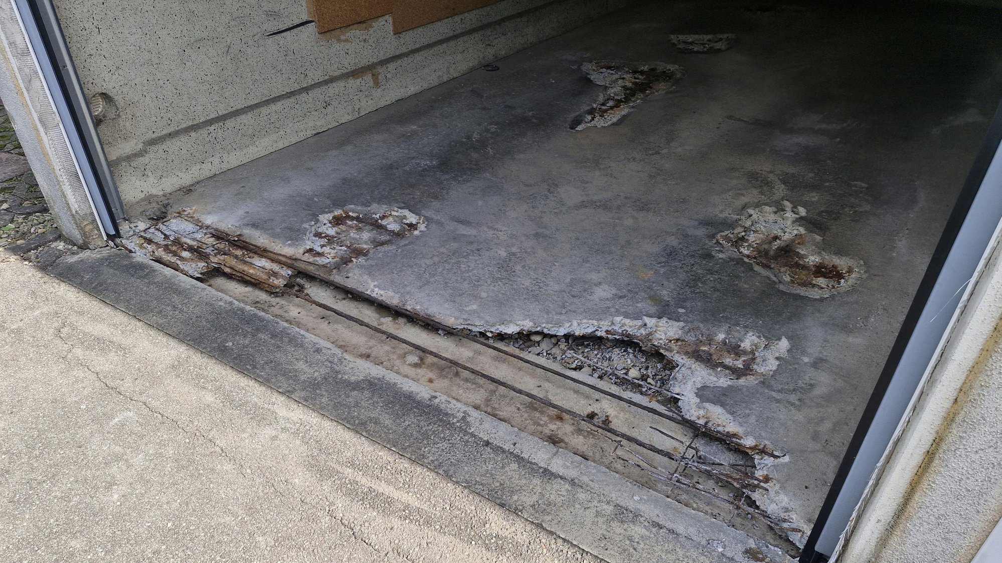 Betonboden mit freiliegendem, verrostetem Bewehrungsstahl und abgeplatzten Stellen am Eingang