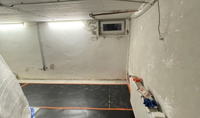 Kellerraum mit unebenen, beschädigten Wänden und schwarzer Folie auf dem Boden