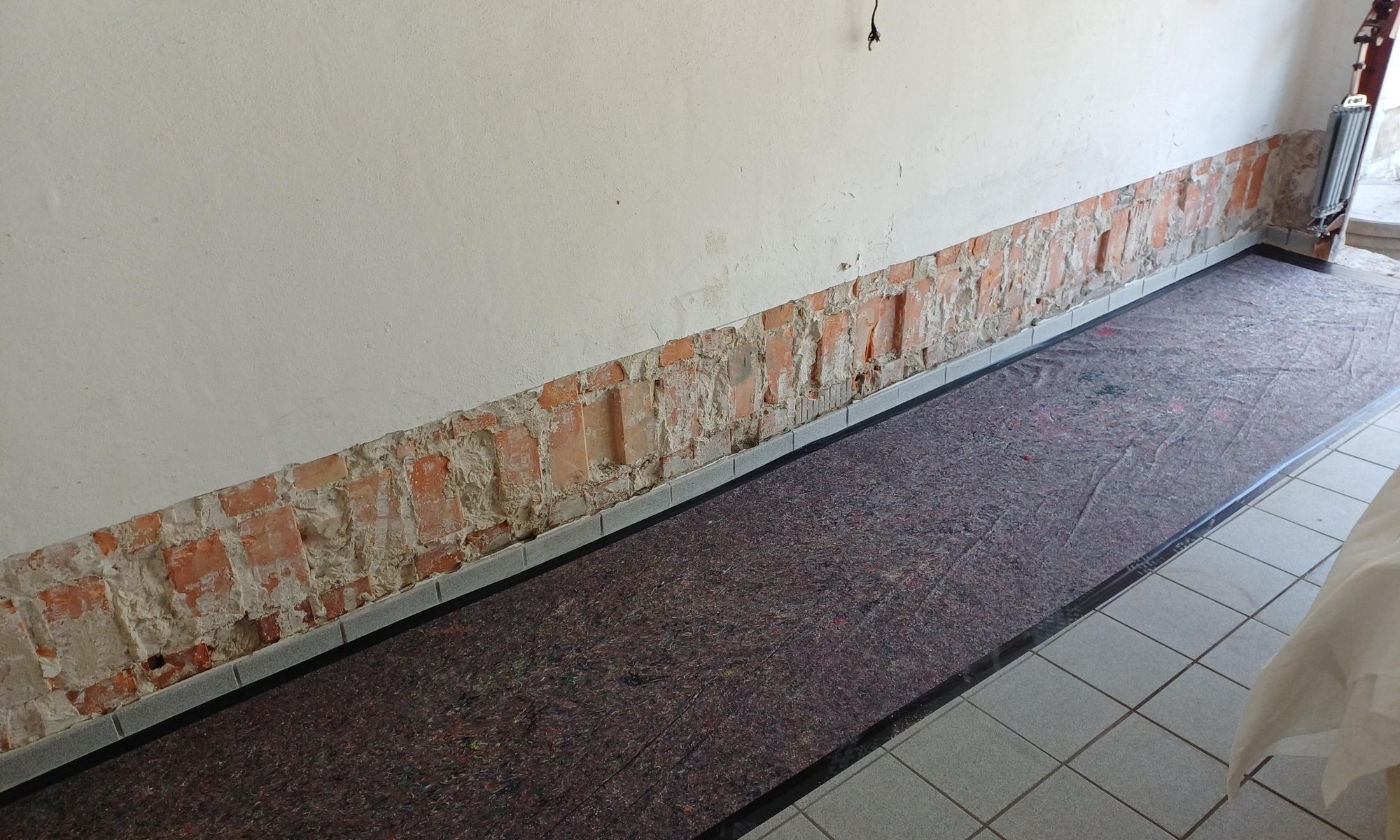 Freigelegte Ziegelreihe am Wandfuß, Boden mit Abdeckvlies und Fliesenrand.