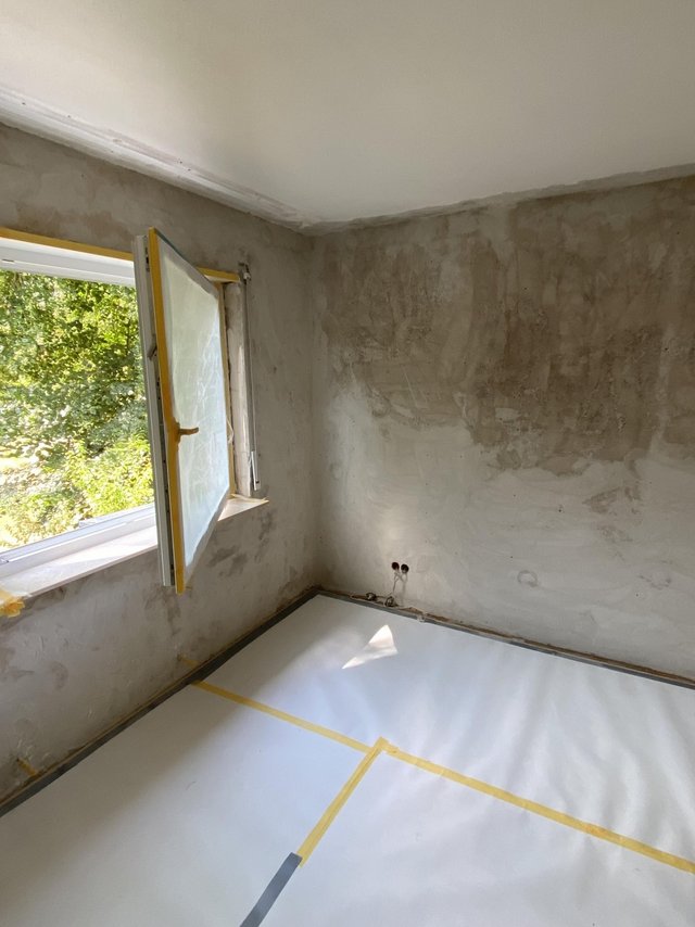Zimmer mit geöffneter Fensterfront, fleckigen Wandflächen und abgeklebtem Boden mit Schutzfolie