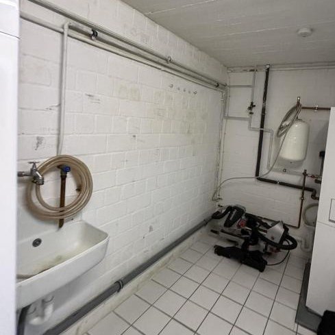 Kellerraum mit Waschbecken, Schlauchaufroller, Leitungen an Ziegelwand und Fliesenboden