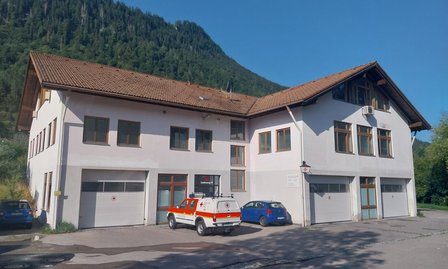 Weißes Gebäude mit Garagentoren, geparkten Autos und bewaldetem Berghang im Hintergrund.