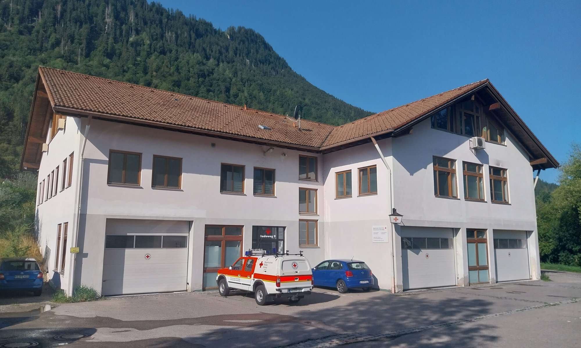 Weißes Gebäude mit Garagentoren, geparkten Autos und bewaldetem Berghang im Hintergrund.