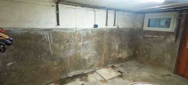Feuchte Kellerwand mit Ausblühungen, Rohrleitungen an der Decke und Fenster.