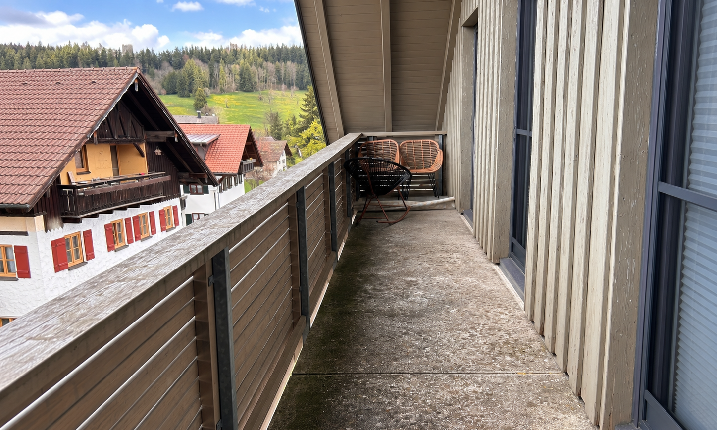 Balkon mit Holzgeländer, Rattanstühlen und Metallstäben, Blick auf Häuser und Wald
