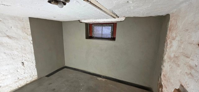 Kellerraum mit grauer Beschichtung an Wand, kleines Fenster, rohe Steinflächen seitlich.