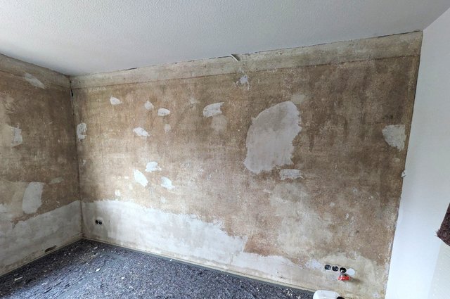 Zimmerwand mit freigelegter brauner Oberfläche und hellen Spachtelstellen, Boden abgedeckt.