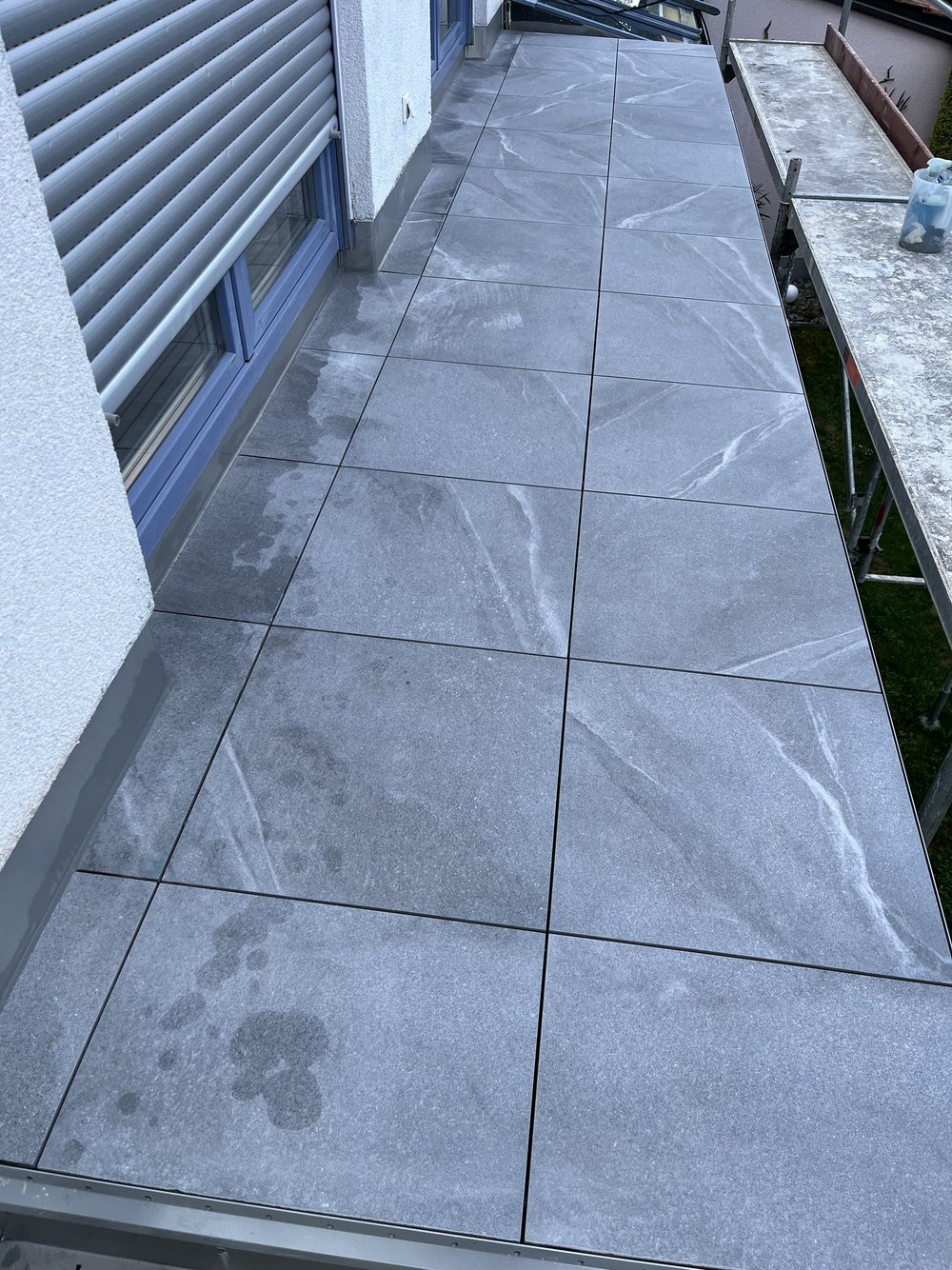Graue Balkonfliesen mit weißen Schlieren, neben Fensterfront und Baugerüst.
