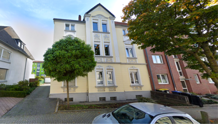 Mehrstöckiges Wohnhaus mit Stuckfassade, Straßenansicht mit Bäumen und geparktem Auto.