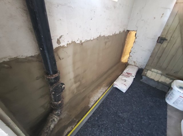 Freigelegte Kellerwand mit grauer Masse und schwarzem Rohr, Nahaufnahme
