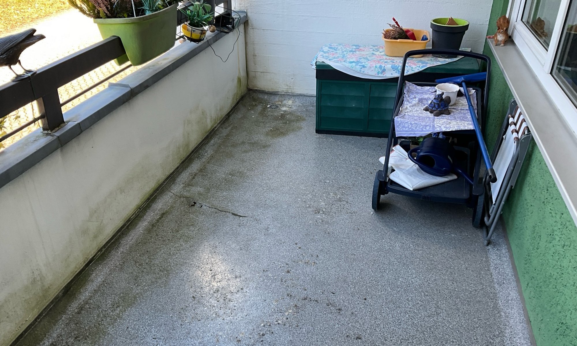 Balkon mit grauem Betonboden, Riss und feuchten Flecken, daneben Pflanzkübel und Gartenwagen