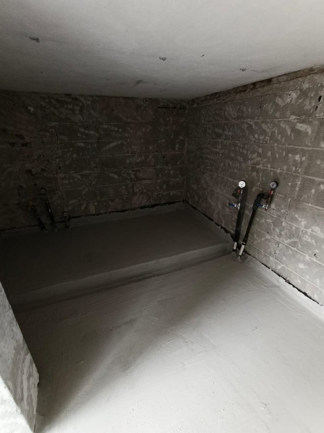 Unfertiger Kellerraum mit Betonwänden, frisch geglättetem Estrich und Rohranschlüssen mit Manometern.
