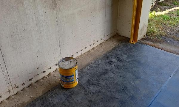 Gelber Eimer auf grauem Boden vor Betonwand mit Bohrlochreihe