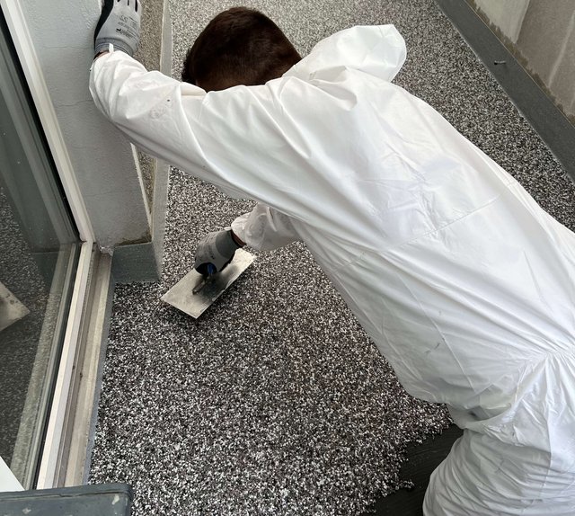 Person im Schutzanzug glättet graue Masse am Balkonboden mit Glättkelle